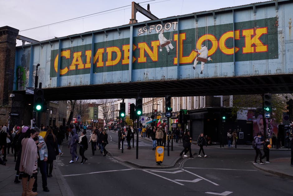 A busy street scene in Camden Town, with a large, weathered metal railway arch bridge overhead displaying the yellow and red logo of Camden Lock; underneath, pedestrians are crossing at a traffic light controlled intersection, some waiting on the pavement and others walking across the road. On the street, there are various cardboard boxes, wrapped furniture items, and packing materials near a moving van, which is partially visible on the right side of the image. The environment is urban with commercial and residential buildings lining the street, and the overall scene depicts a typical day of home relocation or furniture transport activities involving loading or unloading processes, with the presence of moving equipment such as trolley carts and straps operated by Movers Camden in their regular removals service.