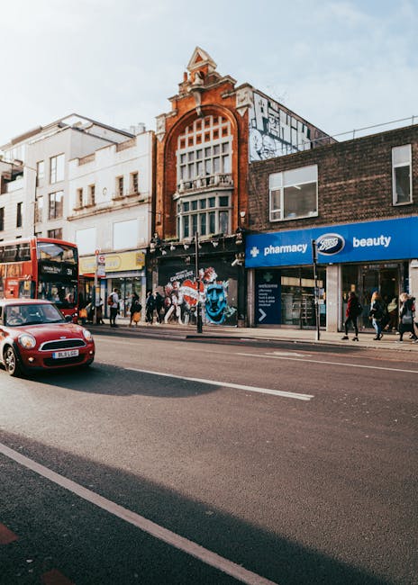 The image shows a busy street scene on Chalk Farm Road NW1 in Camden, featuring a row of commercial buildings with retail shops and offices. In the foreground, a red double-decker bus is seen on the street, along with a red car traveling next to it. Pedestrians are walking along the sidewalk, some entering or exiting the shops, which include a pharmacy in a blue storefront and a beauty store. The buildings have a mix of brick and modern facades, with the central building displaying an ornate brick architectural style, including a large arched window and decorative elements. The street is paved with asphalt, with marked lanes and side pavements. Visible loading and unloading activity occurs in front of the retail stores, where cardboard boxes, plastic wrap, and packing materials are placed near the shop entrances, indicating ongoing home or business relocations. Movers Camden's removal services could be involved in transporting furniture, boxes, and appliances from this busy urban area, supporting efficient packing, loading, and moving logistics amid the vibrant city environment.