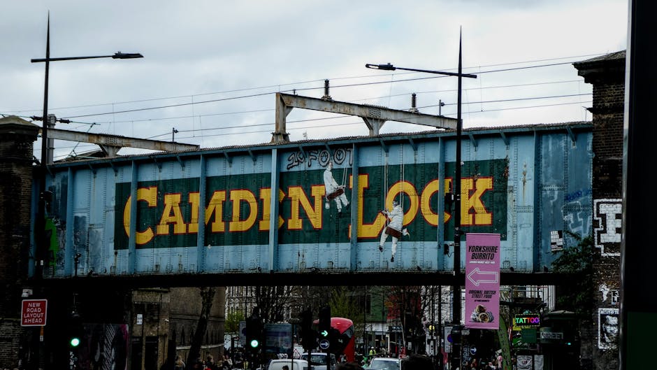 A busy street scene in Camden Town, with a large, weathered metal railway arch bridge overhead displaying the yellow and red logo of Camden Lock; underneath, pedestrians are crossing at a traffic light controlled intersection, some waiting on the pavement and others walking across the road. On the street, there are various cardboard boxes, wrapped furniture items, and packing materials near a moving van, which is partially visible on the right side of the image. The environment is urban with commercial and residential buildings lining the street, and the overall scene depicts a typical day of home relocation or furniture transport activities involving loading or unloading processes, with the presence of moving equipment such as trolley carts and straps operated by Movers Camden in their regular removals service.