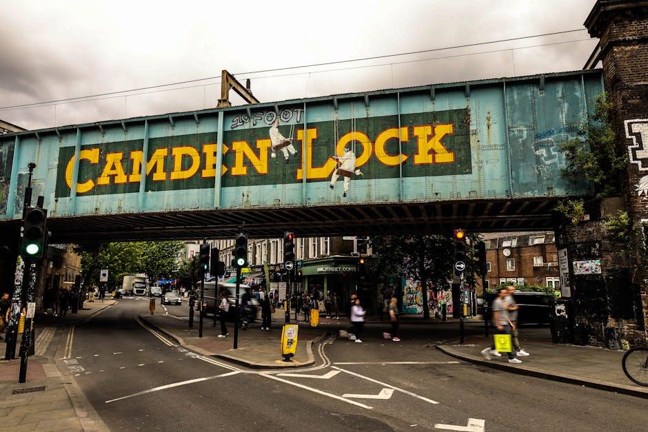 Photograph of a street scene in Camden under the Camden Lock railway bridge, which features large, bold yellow lettering spelling 'CAMDEN LOCK' on a weathered teal metal surface. The bridge spans acro