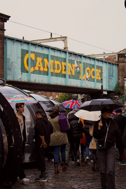 The image shows a crowded street scene near Camden Lock Market, with many pedestrians walking under an old, weathered blue railway bridge bearing the yellow and orange signage of 'CANDEN LOCK'. Some people are holding umbrellas in various colors, likely due to rain, while others are dressed in dark clothing suitable for wet weather. Several vehicles, including black vans, are parked or positioned nearby, suggesting a busy area for home relocation or furniture transport. The pavement underneath is cobbled, and the overall environment appears to be an urban setting close to the Roundhouse in Camden, with a mix of people engaged in moving or delivering items. This scene reflects the busy logistics involved in house removals, fitting with professional moving services such as those offered by Movers Camden, particularly in central London locations like Camden Lock Market and the Roundhouse area.