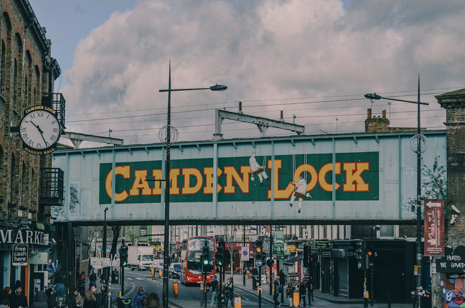 A large, painted metal sign with the words 'Camden Lock' in bold yellow letters on a green background is suspended above a busy street scene in Camden, London. The sign is attached to a metal framework spanning across the road, with visible support beams and hanging electrical wires. Surrounding the sign are street lamps on tall poles and a clock mounted to a building on the left, showing approximately 2:44. Below, there is a mix of pedestrians walking along shopfronts and street vendors, with some individuals wearing backpacks and carrying shopping bags. A red double-decker bus is visible moving along the street, and various vehicles, including cars and smaller bikes, are present. The scene features diverse people, some waiting at crosswalks, among shop entrances and street signage. This environment exemplifies the busy, urban atmosphere typical of a home relocation or furniture transport process in Camden, with visible move-related activity and signage, supporting the context of house removals and moving services by Movers Camden. Natural daylight illuminates the scene with some cloud cover overhead.