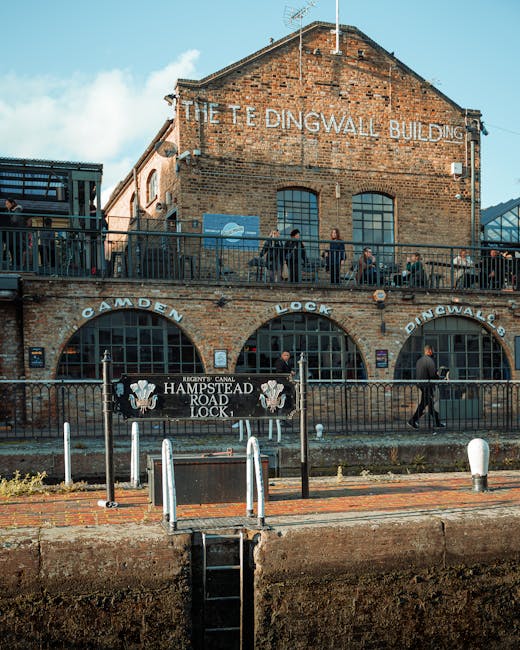 The image shows a crowded street scene near Camden Lock Market, with many pedestrians walking under an old, weathered blue railway bridge bearing the yellow and orange signage of 'CANDEN LOCK'. Some people are holding umbrellas in various colors, likely due to rain, while others are dressed in dark clothing suitable for wet weather. Several vehicles, including black vans, are parked or positioned nearby, suggesting a busy area for home relocation or furniture transport. The pavement underneath is cobbled, and the overall environment appears to be an urban setting close to the Roundhouse in Camden, with a mix of people engaged in moving or delivering items. This scene reflects the busy logistics involved in house removals, fitting with professional moving services such as those offered by Movers Camden, particularly in central London locations like Camden Lock Market and the Roundhouse area.